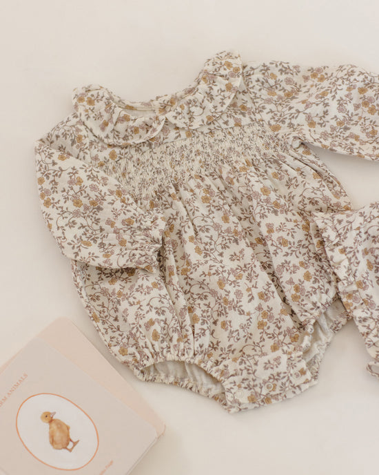 A baby long-sleeve romper with a smocked chest and ruffled collar, featuring a delicate beige and brown floral print on a cream background, styled flat-lay on a light cream surface next to a book.