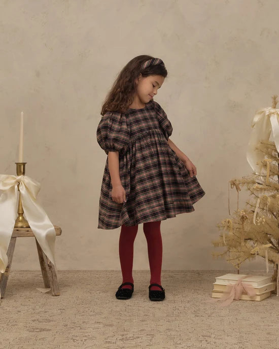 A young girl stands in a studio setting wearing a dark brown and black plaid dress with puff sleeves, red tights, and black shoes. The background features a textured beige wall, a small decorated Christmas tree with gold ornaments, and a rustic wooden stool with a white ribbon and candle.