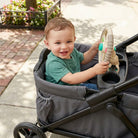 Child playing with a toy steering wheel in a stroller on a sidewalk.