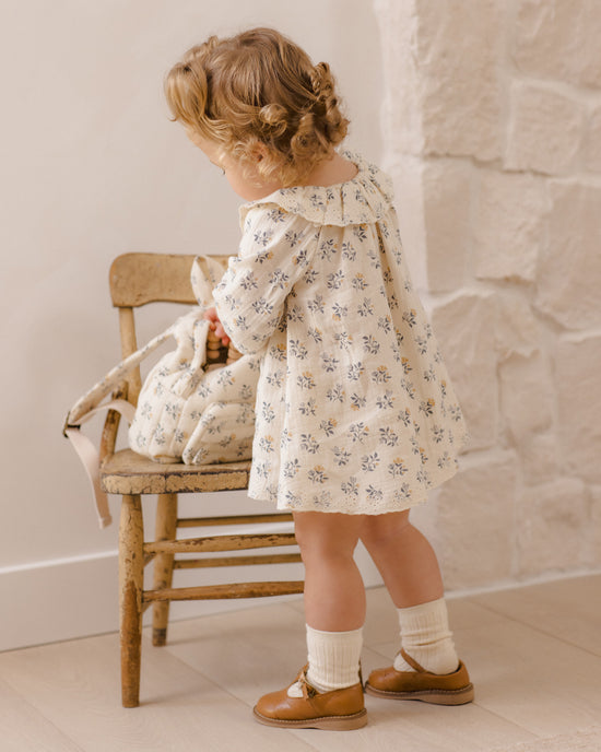 A toddler girl stands beside a rustic wooden chair in a warm indoor setting, wearing a cream long-sleeve dress with a small blue floral print, white socks, and brown shoes.