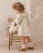 A toddler girl stands beside a rustic wooden chair in a warm indoor setting, wearing a cream long-sleeve dress with a small blue floral print, white socks, and brown shoes.