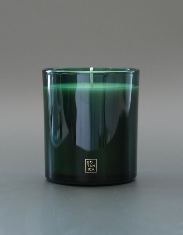 A dark green glass jar candle with a single white wick, featuring a small gold square logo on the front that reads 'BOTANICA'. The candle is photographed against a neutral gray background.