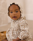 A baby wearing a cream-colored floral smocked romper and matching bonnet, sitting next to a woven basket.