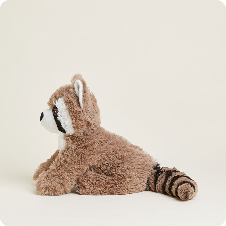A side-profile studio product photo of a plush Raccoon by Warmies lying down against a white background, highlighting its soft brown fur, white-trimmed ear, and striped tail.