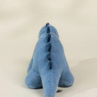 A blue plush dinosaur toy shown from the back, featuring a soft fuzzy texture and a darker blue stitched spine, set against a plain light beige background.