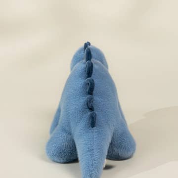 A blue plush dinosaur toy shown from the back, featuring a soft fuzzy texture and a darker blue stitched spine, set against a plain light beige background.
