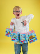 A young boy with glasses and an open mouth expression holds four colorful 'Puzzle' jigsaw puzzle gift boxes against a solid yellow background.