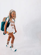 A young girl walking on a white studio background wearing a white graphic t-shirt that says 'KIND GIRL', white shorts, patterned socks, and carrying a teal and brown backpack.