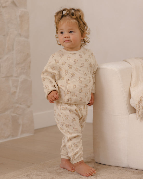 A toddler stands barefoot indoors wearing a cream-colored two-piece pajama set with a small beige floral pattern. The set includes a long-sleeve top and matching pants.