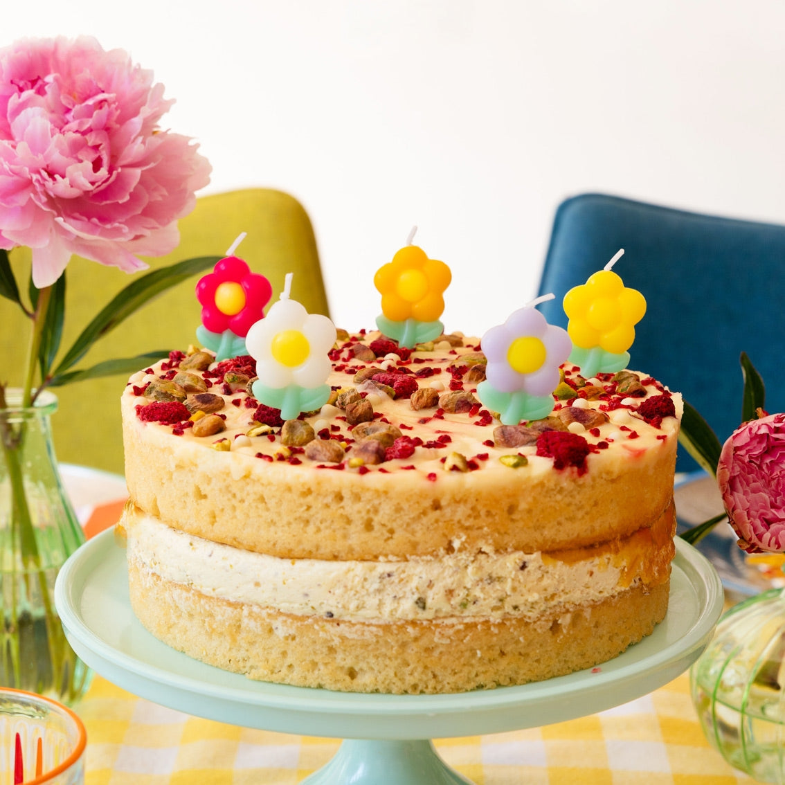 Two-layer cake with colorful candles on a green cake stand, surrounded by flowers and a yellow chair.