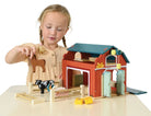 A young girl with blonde braids, wearing a short-sleeved yellow dress, plays with the Wooden Big Red Barn Playset by Mentari, holding a wooden toy horse near the open barn doors and fenced enclosure.
