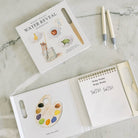 A children's water painting kit shown open on a light marble surface, with the illustrated box, a wooden palette tool, two white paint pens, and a spiral-bound notebook page that reads 'Whiny Washy Wasty Wry Wry SWISH SWISH'.