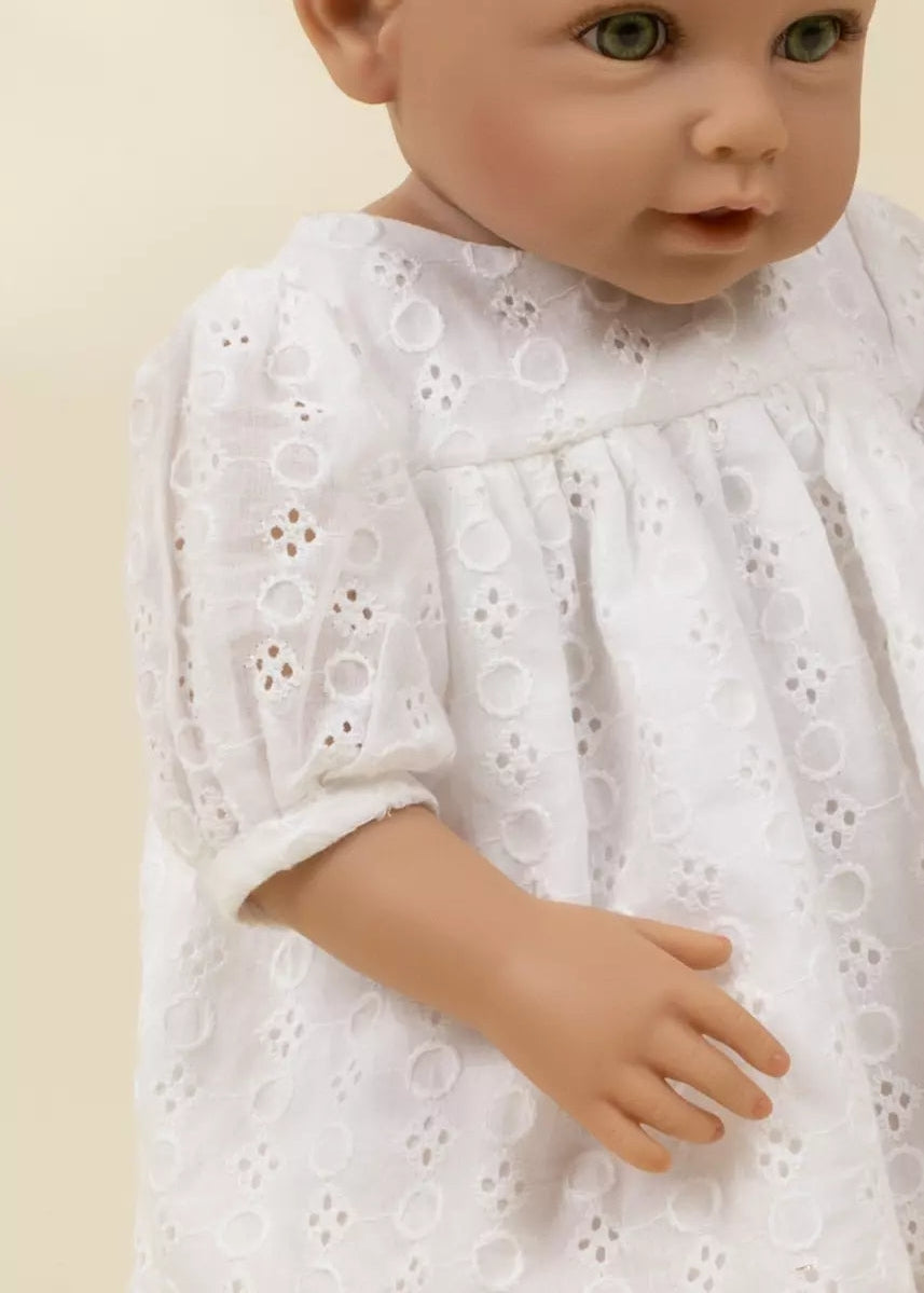 A close-up of a doll or toy baby wearing a white eyelet lace dress against a plain light beige background.