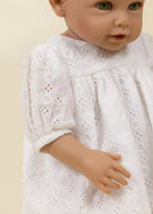 A close-up of a doll or toy baby wearing a white eyelet lace dress against a plain light beige background.