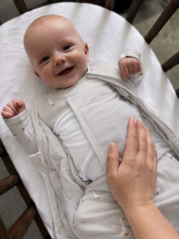 A smiling baby wearing a white long-sleeve sleep sack with tiny brown animal prints, lying in a wooden bassinet while an adult hand gently touches the baby's back.