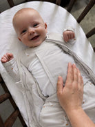 A smiling baby wearing a white long-sleeve sleep sack with tiny brown animal prints, lying in a wooden bassinet while an adult hand gently touches the baby's back.
