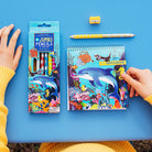 Children's art supplies including pencils, a sketchbook with an ocean design, and a sharpener on a blue background.