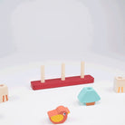 Wooden Pet Stacker toy by Mentari featuring a red base with pegs, with an orange and yellow bird piece lying in the foreground and a disassembled pastel blue block nearby against a plain white background.