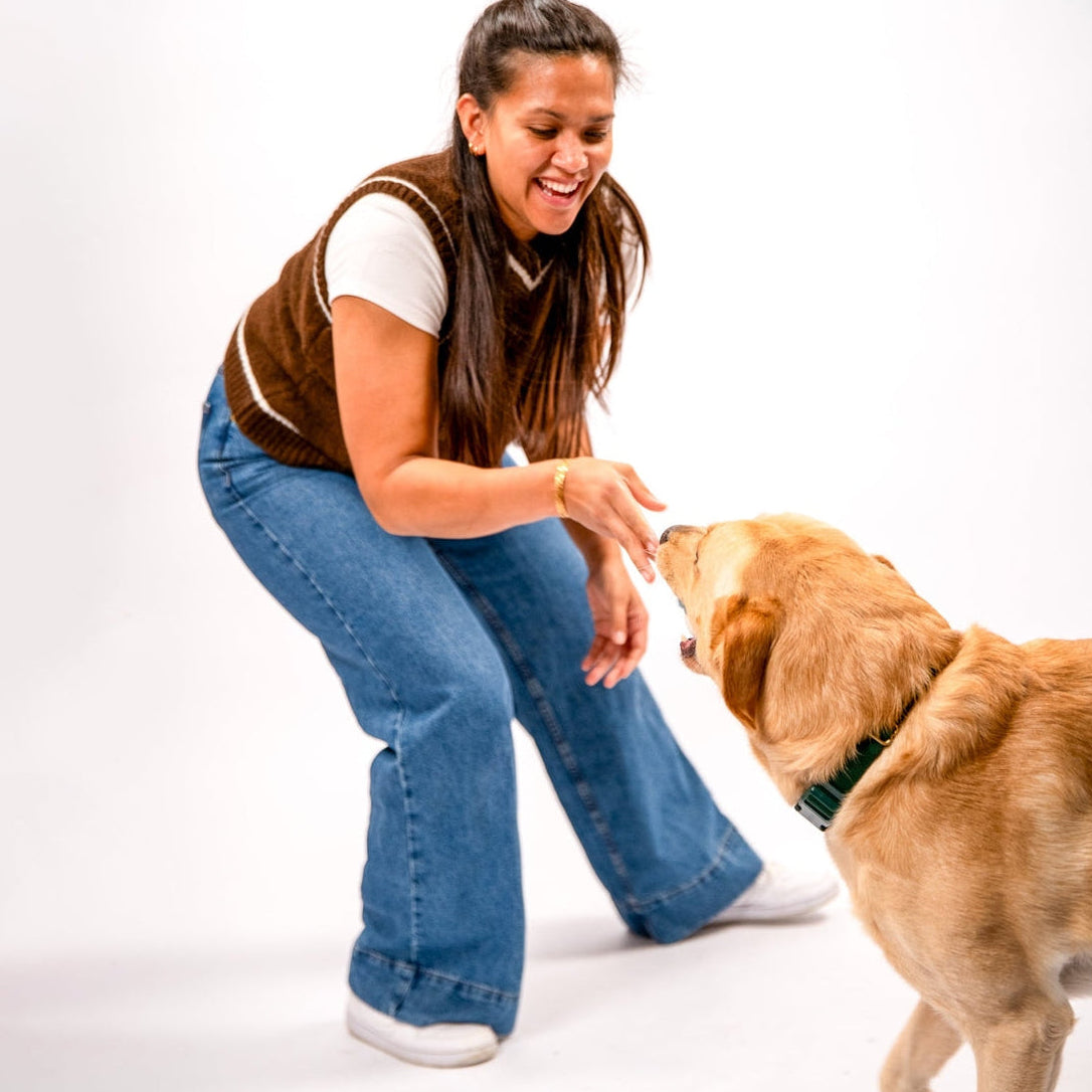 A woman in a brown vest and blue jeans crouches down to greet a golden retriever dog wearing a green collar, set against a plain white studio background.