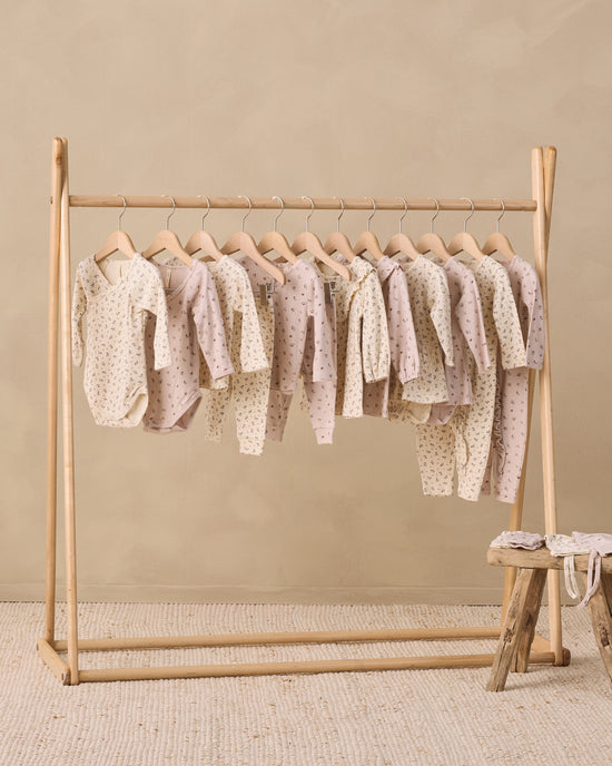 A wooden clothing rack holding multiple baby long-sleeve bodysuits and onesies in soft blush, cream, and beige tones with small polka dots, set against a warm beige wall and textured rug.