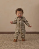 A toddler with curly brown hair stands on a patterned rug wearing a beige and olive green plaid button-front romper and brown moccasin-style shoes, against a plain beige wall.