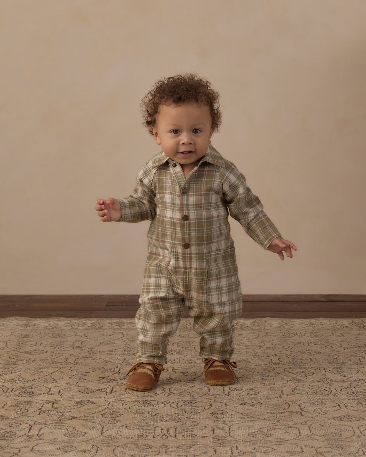 A toddler with curly brown hair stands on a patterned rug wearing a beige and olive green plaid button-front romper and brown moccasin-style shoes, against a plain beige wall.