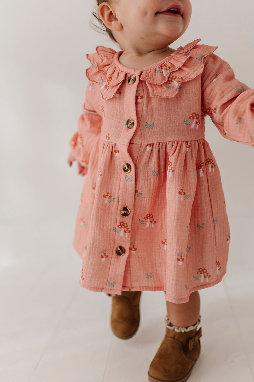 A toddler wearing a dusty pink long-sleeve dress with a ruffled collar and small mushroom prints, paired with brown boots, photographed against a plain white background.