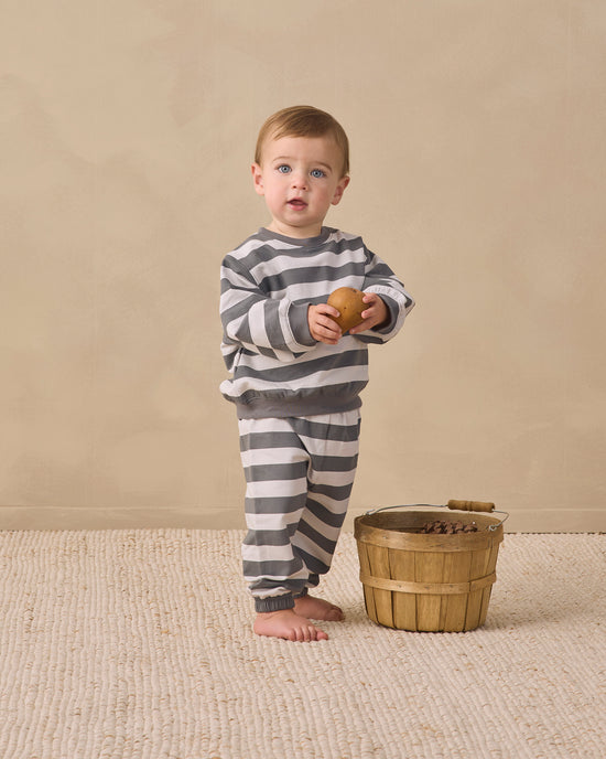 A toddler stands barefoot on a textured beige rug against a warm beige wall, wearing a gray and white striped long-sleeve top and matching pants. The child holds a small brown apple and stands next to a wooden basket.