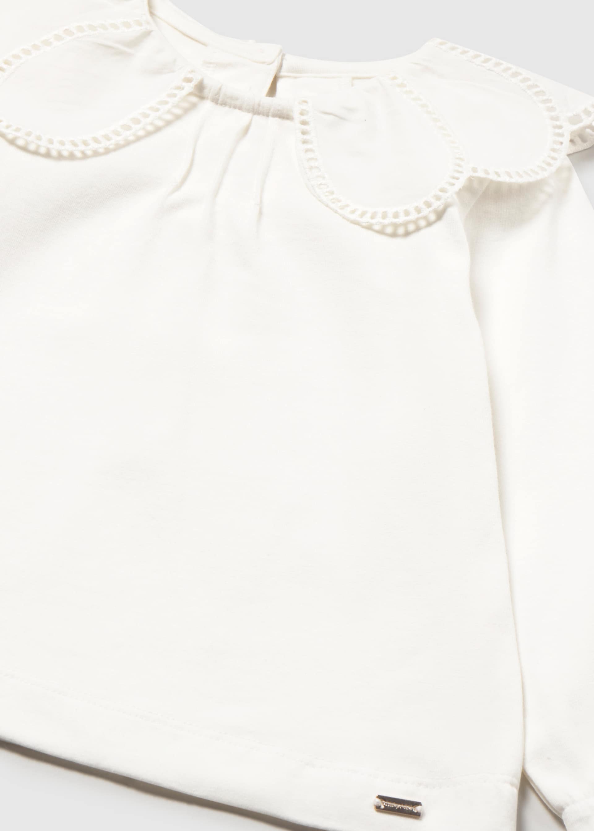Close-up of a white long-sleeve blouse with a large decorative ruffled collar and a small metallic brand tag near the lower hem.