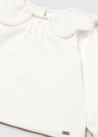 Close-up of a white long-sleeve blouse with a large decorative ruffled collar and a small metallic brand tag near the lower hem.