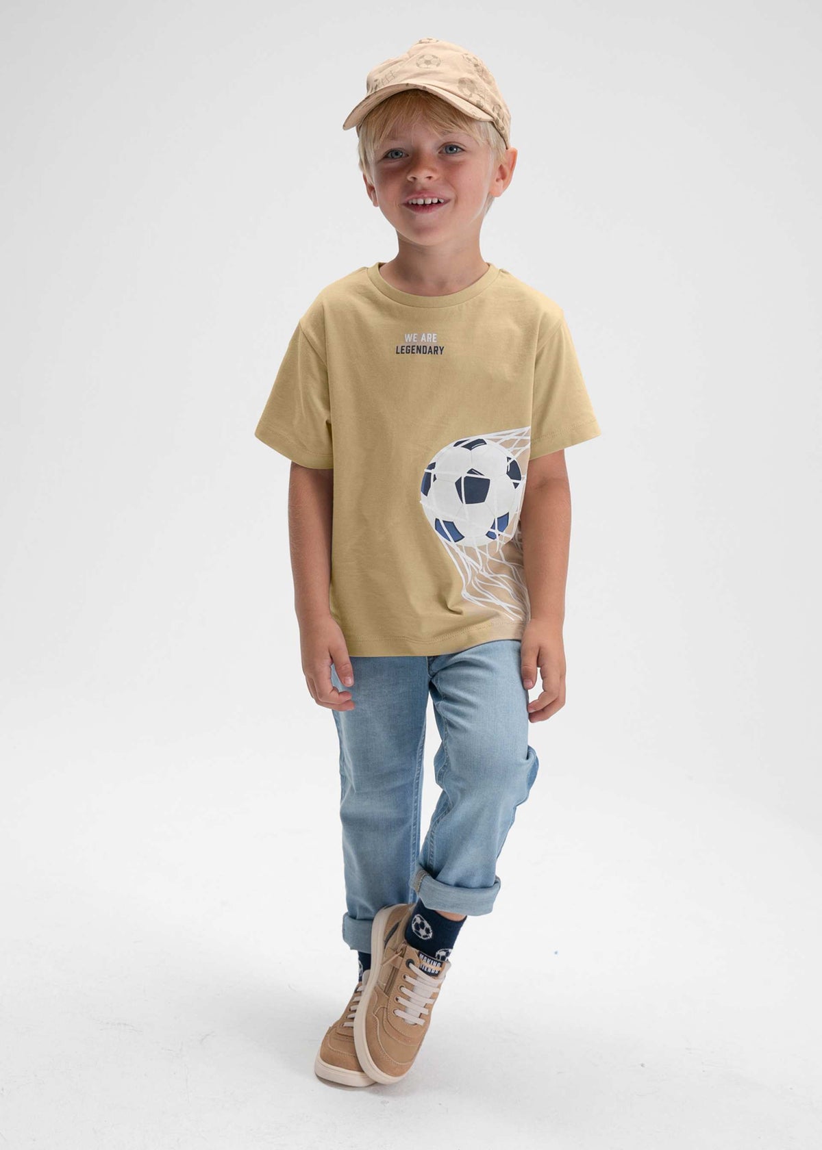 Child wearing a beige t-shirt with a graphic design, light blue jeans, and a cap on a white background