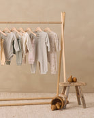 A wooden clothing rack holding several baby long-sleeve onesies in soft neutral tones, including striped, solid pale pink, and floral prints. A small rustic stool with pears sits beside the rack against a warm beige wall and textured rug.