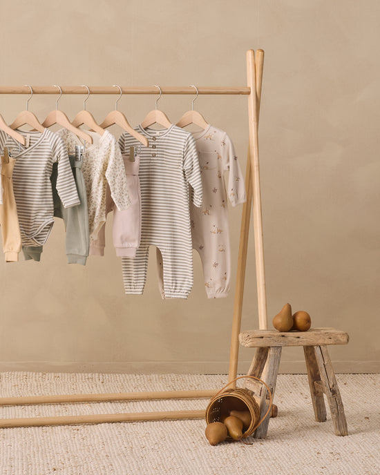 A wooden clothing rack holding several baby long-sleeve onesies in soft neutral tones, including striped, solid pale pink, and floral prints. A small rustic stool with pears sits beside the rack against a warm beige wall and textured rug.