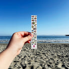 A hand holds a vertical fish-count chart in front of a sandy beach and blue ocean under a clear blue sky. The chart has rows of illustrated fish and numbers, with a pink ribbon logo near the bottom.
