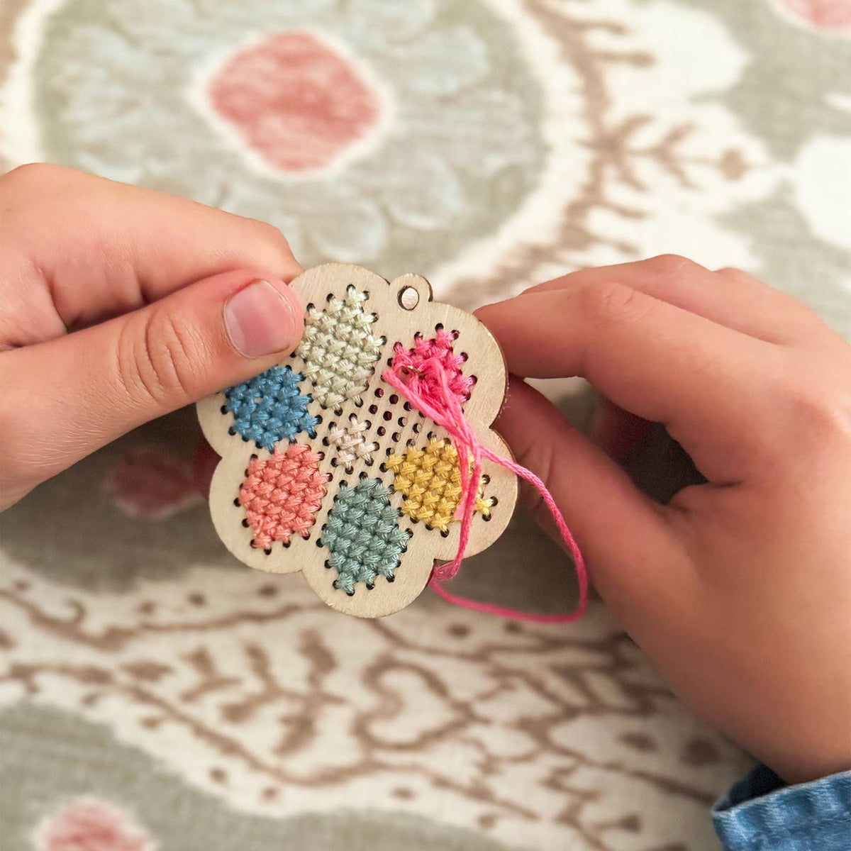 Hand holding a small embroidered wooden ornament with colorful patterns on a patterned fabric background.