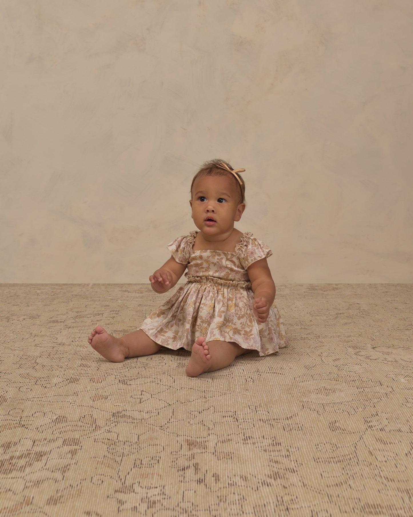 A baby girl sitting on a textured beige rug against a neutral beige wall, wearing a short-sleeve cream dress with a yellow floral pattern and a matching yellow bow headband.