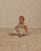 A baby girl sitting on a textured beige rug against a neutral beige wall, wearing a short-sleeve cream dress with a yellow floral pattern and a matching yellow bow headband.