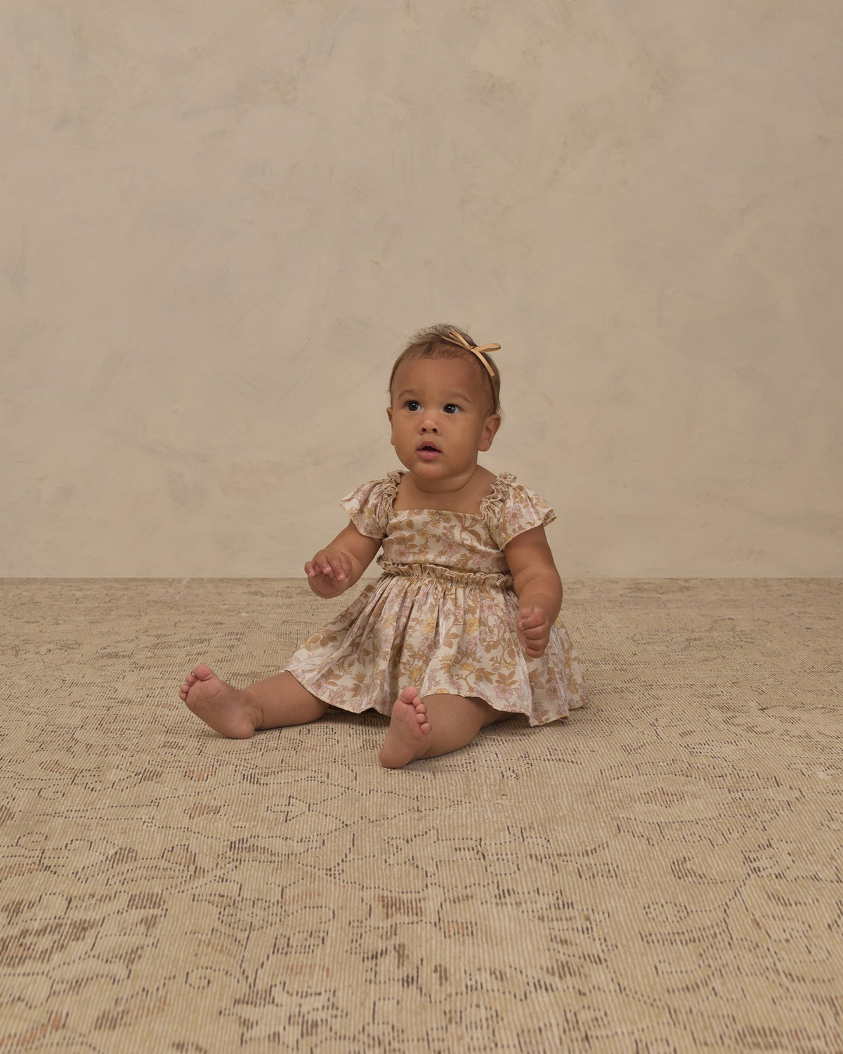 A baby girl sitting on a textured beige rug against a neutral beige wall, wearing a short-sleeve cream dress with a yellow floral pattern and a matching yellow bow headband.