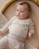 A baby lying on a woven bassinet, wearing a cream and beige floral long-sleeve wrap top with matching beige pants and a light gray bow headband.