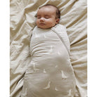 A sleeping newborn baby swaddled in a light beige muslin swaddle blanket with a white goose pattern, lying on a matching beige sheet.