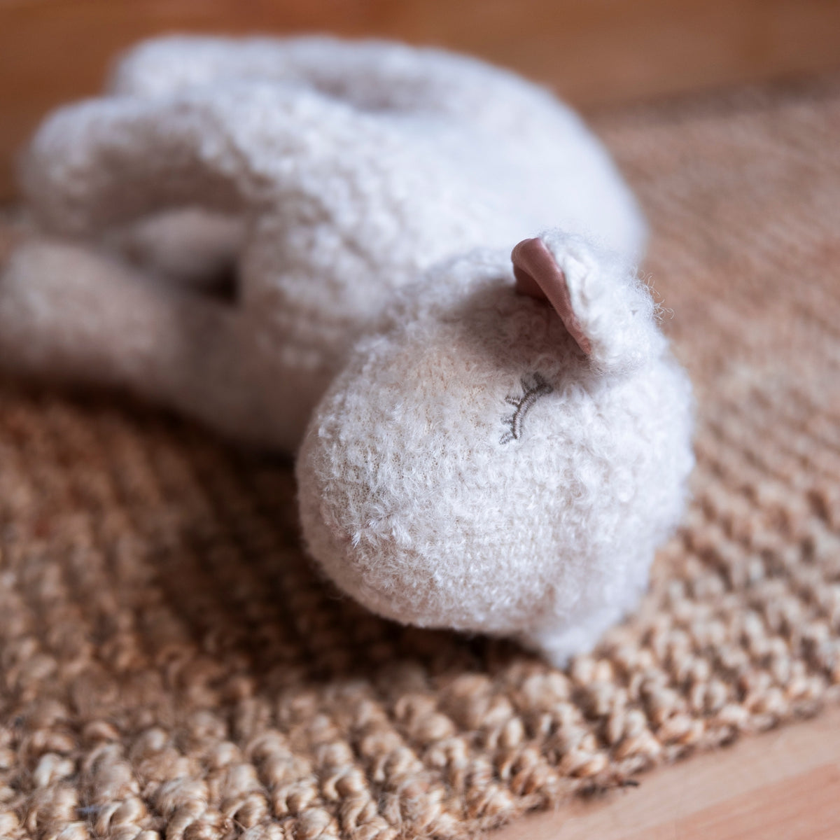 A soft, fuzzy white stuffed lamb with a small pink ear and embroidered closed eyes, resting on a woven jute mat.