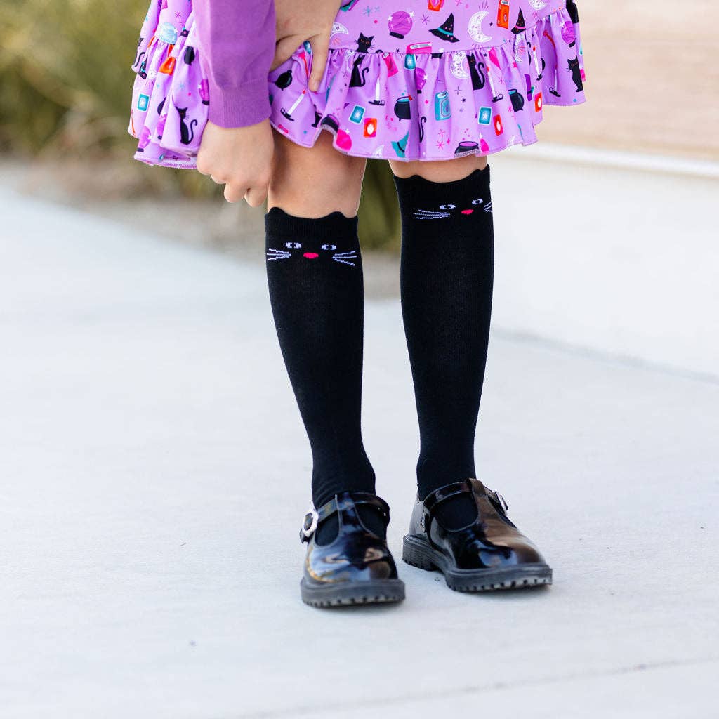 Child wearing a dress with cat print, black knee-high socks with cat faces, and black shoes on a light background.