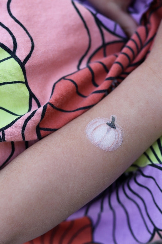 A close-up of a person's forearm featuring a small white pumpkin temporary tattoo with a gray stem, resting over a colorful fabric background with pink, purple, and green patterns.