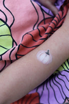 A close-up of a person's forearm featuring a small white pumpkin temporary tattoo with a gray stem, resting over a colorful fabric background with pink, purple, and green patterns.