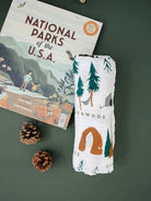 Folded blanket with nature-themed design next to a book titled 'National Parks of the U.S.A.' on a green background