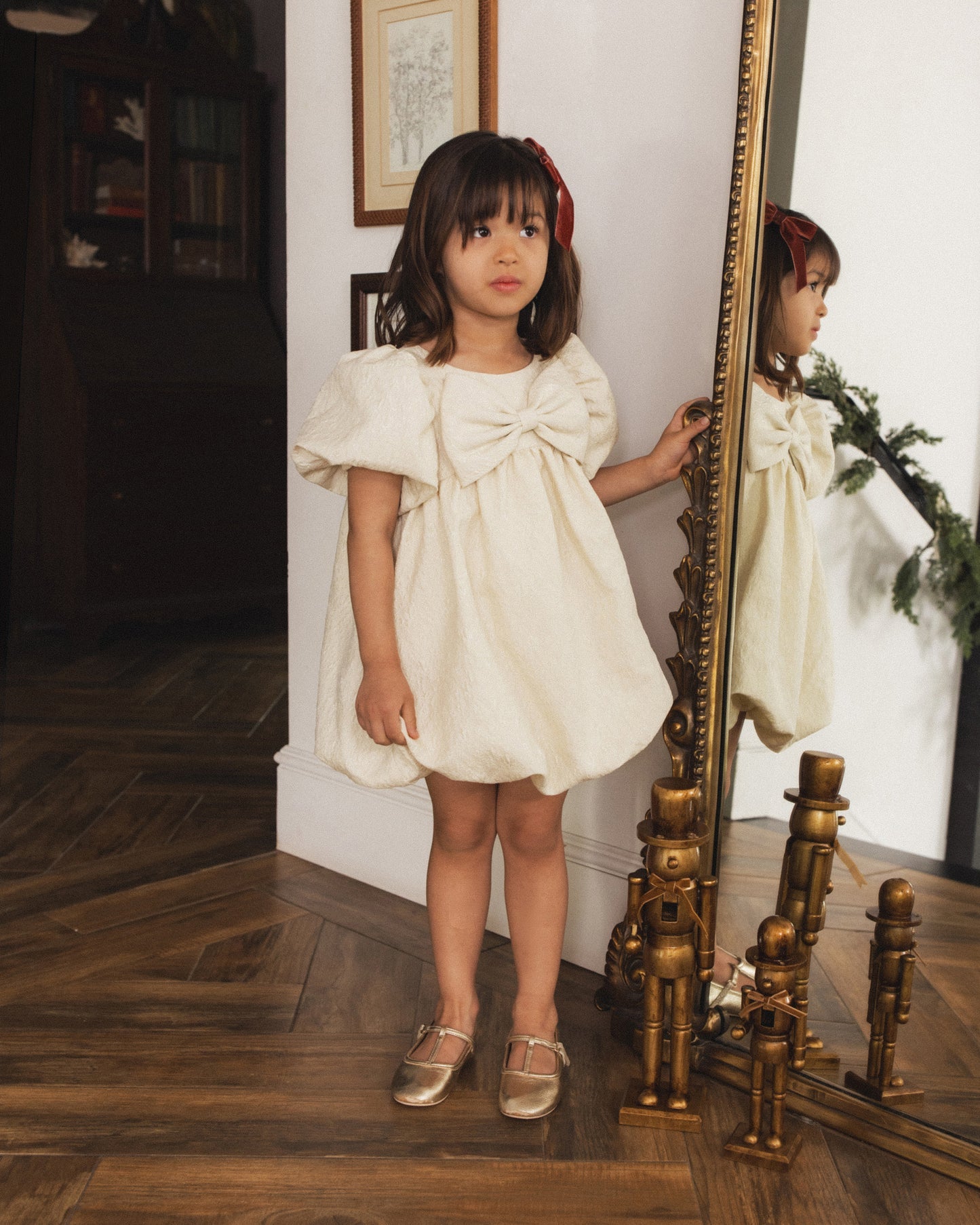 A young girl with dark brown hair wearing a cream-colored dress with a large bow on the chest, standing in a room with a wooden floor and an ornate gold mirror.