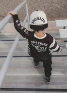 A child wearing a black and white 'Speedster' motocross jersey, black pants, and a white/black trucker hat walks down outdoor wooden steps.