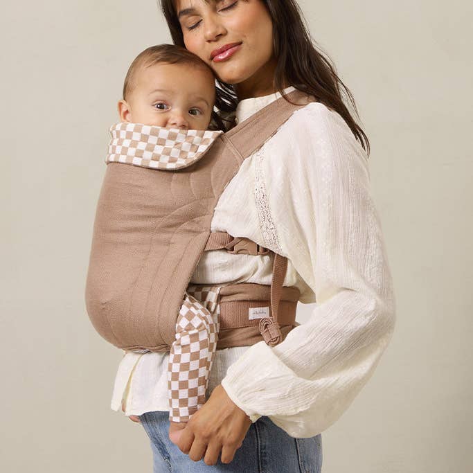 Woman holding a baby in a brown baby carrier against a plain background