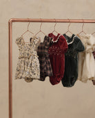 Five baby dresses and smock suits in various patterns and colors hanging on a copper-colored clothing rack against a textured beige wall.
