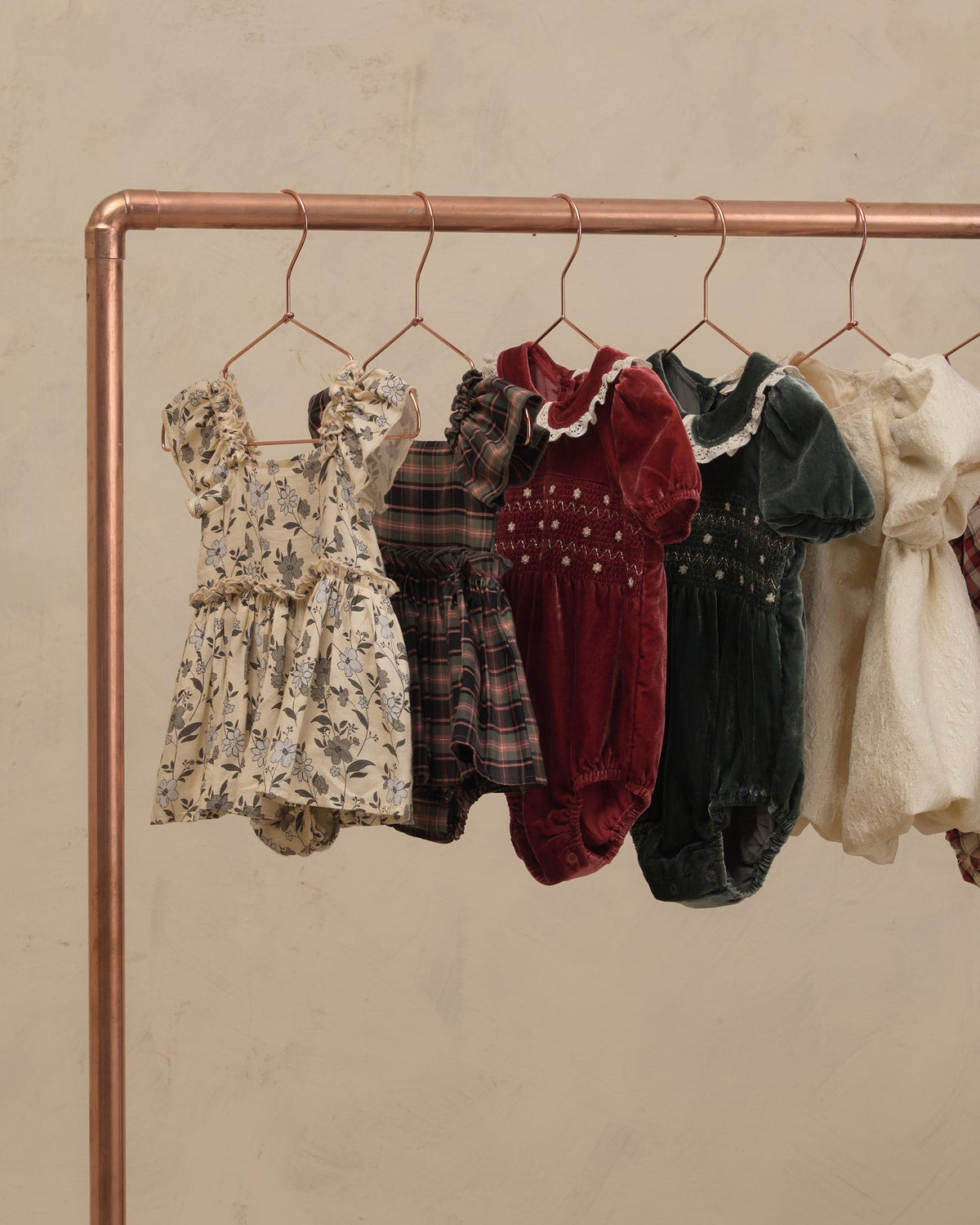 Five baby dresses and smock suits in various patterns and colors hanging on a copper-colored clothing rack against a textured beige wall.
